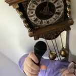 At the top of the photo is an antique wall clock, ornately decorated with brass weights hanging down. Cat stands below in a purple jumper, looking up at the clock and holding up a microphone.