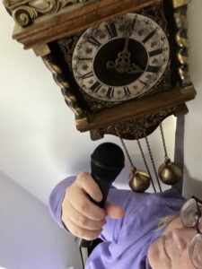 At the top of the photo is an antique wall clock, ornately decorated with brass weights hanging down. Cat stands below in a purple jumper, looking up at the clock and holding up a microphone.